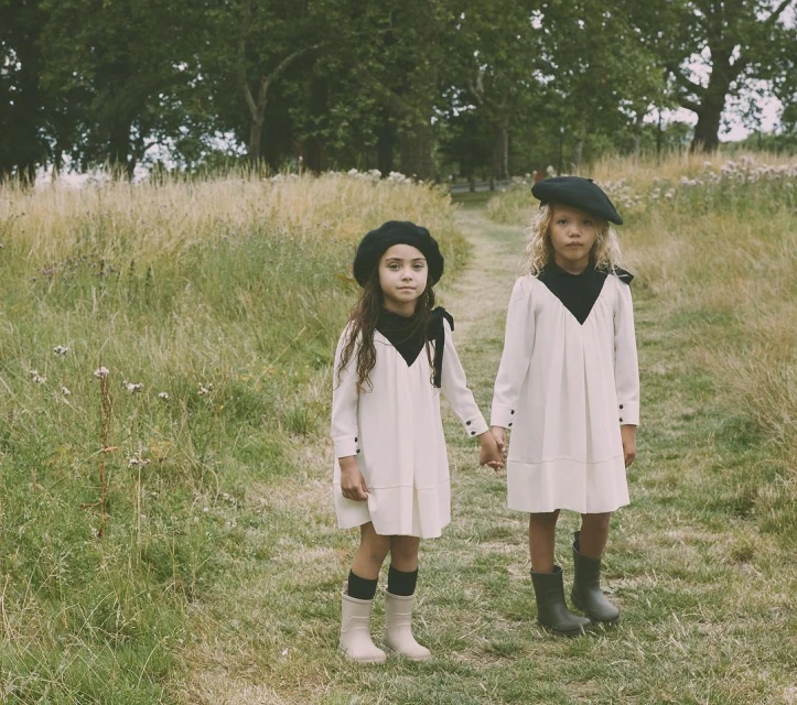 Two young girls in matching dresses and berets hold hands in a grassy field, with a path and trees in the background, creating a vintage feel.