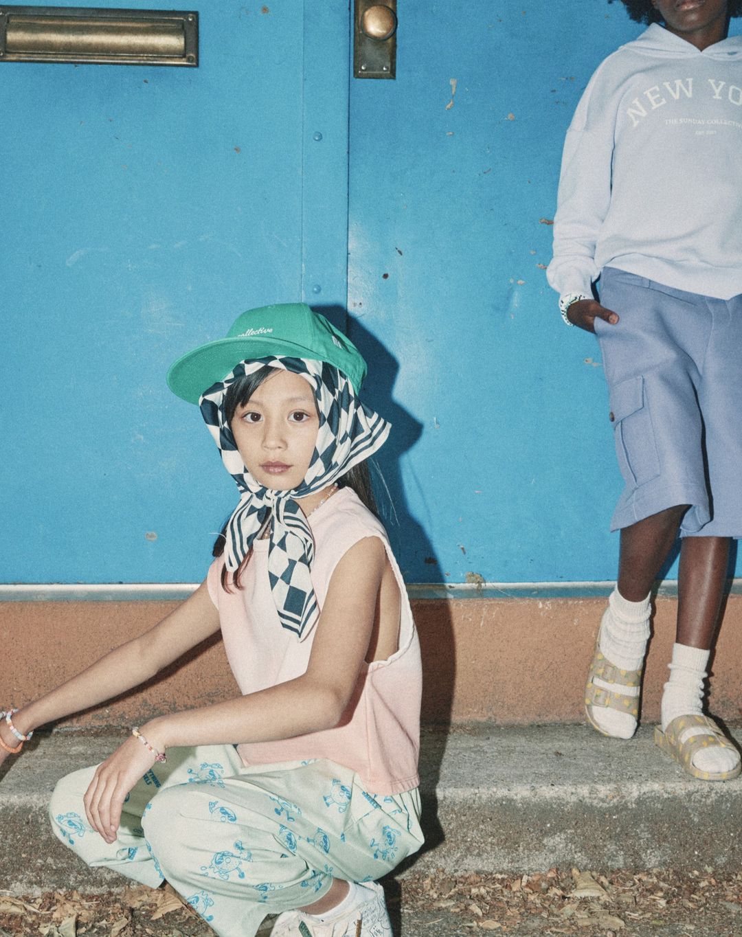 Two young people pose in front of a blue door wearing streetwear including a hoodie, cap, shorts, pants, kerchief and sandals.