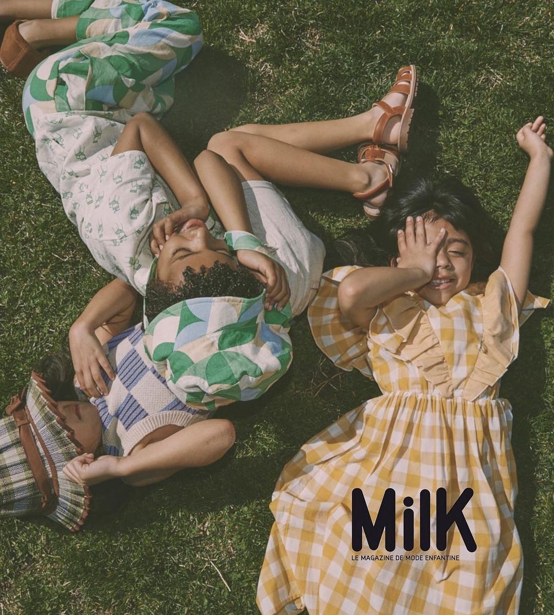 Three smiling children lie on grass in sunlight wearing colourful vintage-style clothes; Milk magazine logo visible at the lower right.