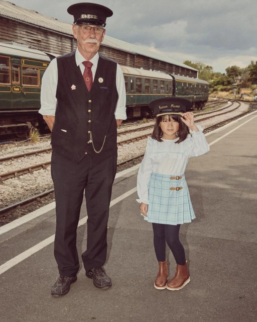 Mature train conductor poses with a child in matching conductor hat on a railway platform with passenger cars in the background.