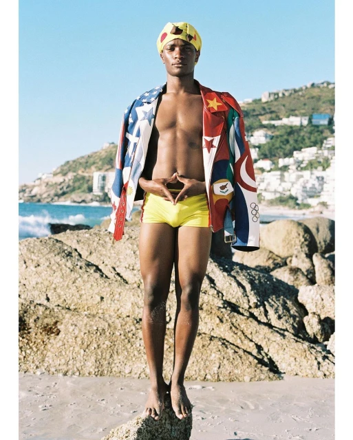 An adult male stands on a beach with rocks in the background, wearing a yellow swimsuit and jacket with global flag designs.