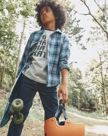 Young boy with curly hair holding a skateboard and orange helmet standing outdoors on a path framed by trees