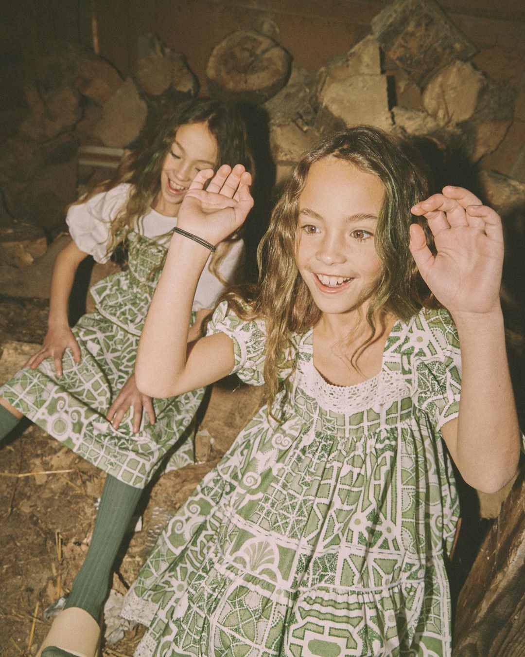 Two smiling girls in matching patterned dresses pose indoors against a wood background, lit with soft, warm lighting in a candid moment.