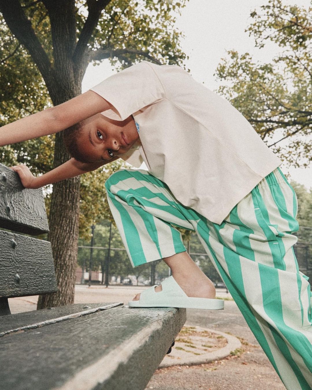 Young person in casual wear poses on a park bench in summer. Mint green and white striped pants and matching slides are visible.