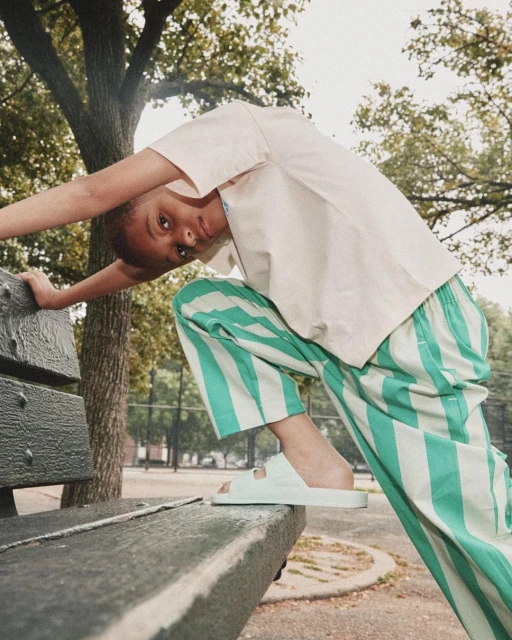 Young person in casual wear poses on a park bench in summer. Mint green and white striped pants and matching slides are visible.