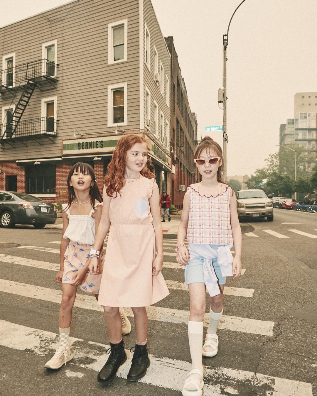 Three young girls in stylish outfits walk across a crosswalk in an urban setting, with buildings and parked cars in the background.