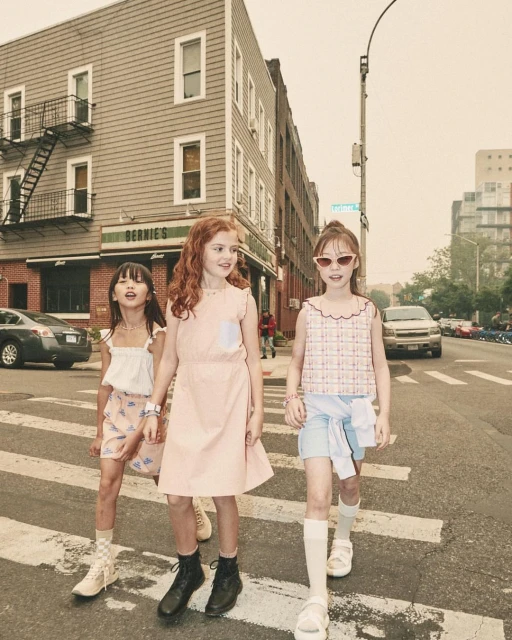 Three young girls in stylish outfits walk across a crosswalk in an urban setting, with buildings and parked cars in the background.