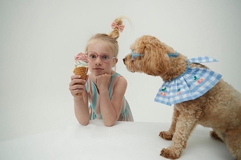 A girl in pink glasses holds an ice cream cone while a poodle wearing glasses and a blue collar looks on in a bright studio shot.