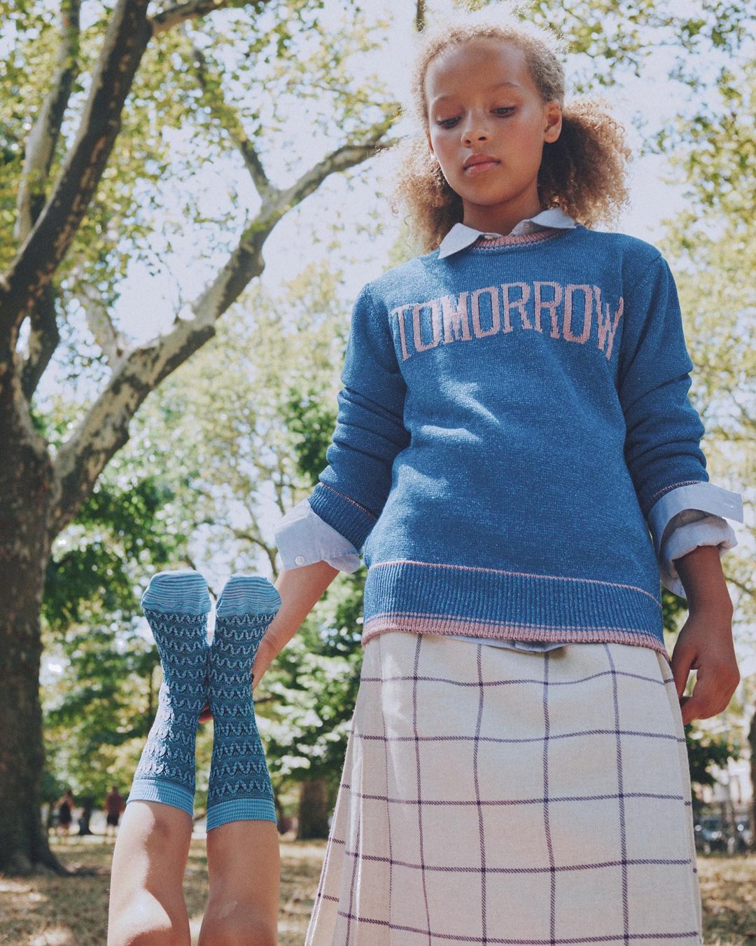 Young girl in a blue 'Tomorrow' sweater and plaid skirt stands outdoors while a pair of legs in patterned socks point skyward.