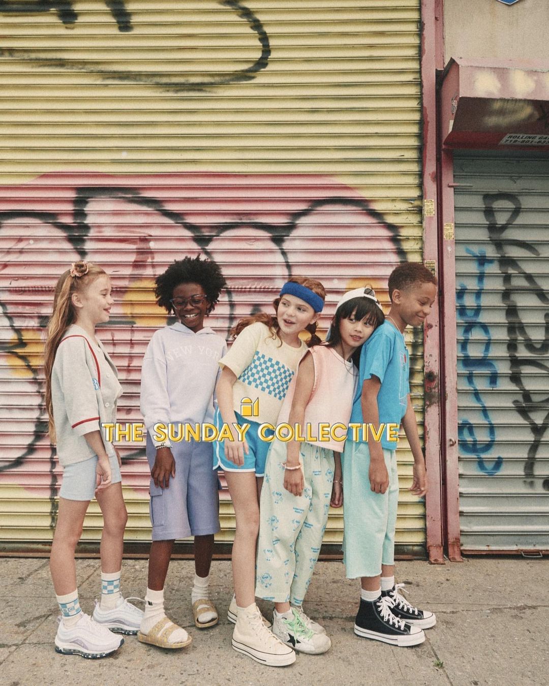 Group of six children stand in front of a graffitied wall wearing casual clothes and sneakers, showcasing youth fashion.