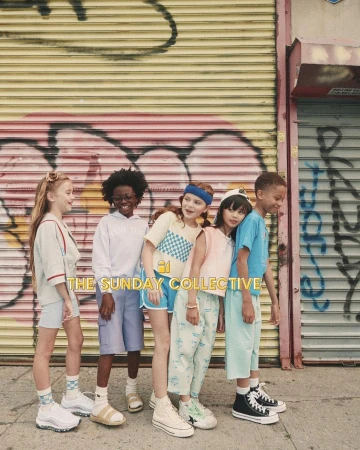 Group of six children stand in front of a graffitied wall wearing casual clothes and sneakers, showcasing youth fashion.