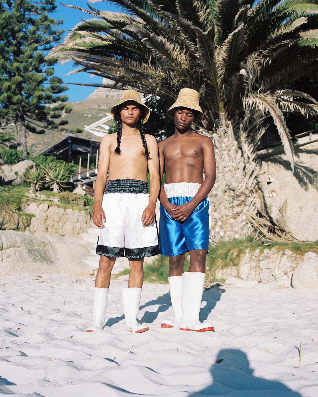 Two shirtless men in boxing shorts and straw hats stand on a sandy beach with palm trees and a distant mountain in the background.