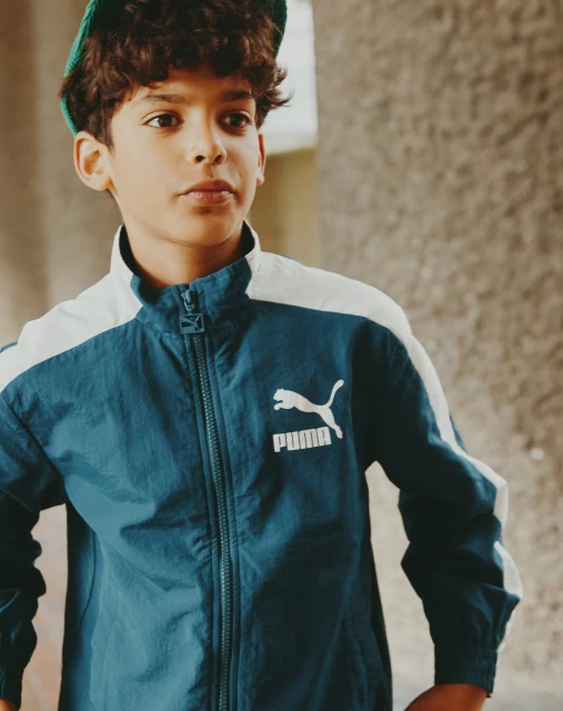 Boy in teal Puma track jacket and green hat poses indoors near neutral-toned wall, lit naturally for a fashion or lifestyle shoot.