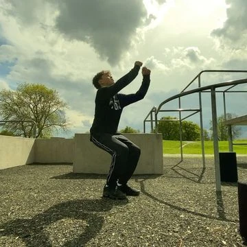 Young man in athletic wear crouches on a textured surface in an outdoor parkour setting with metal bars, green grass, and clouds above.