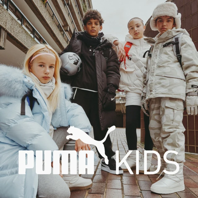 A group of young children in winter clothing pose in an urban outdoor setting with a modern concrete building and the Puma Kids logo.