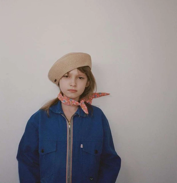 Portrait of a young girl in a blue jacket and tan beret against a neutral wall, wearing a floral neck scarf for a stylish look.