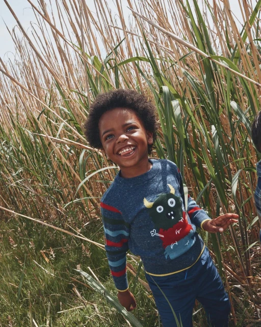 A happy African-American child in a field of tall grasses wears a blue sweater with a monster graphic, smiling with teeth showing.