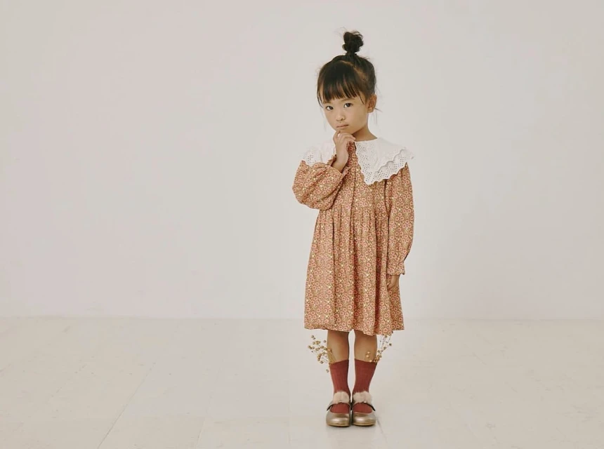 A young girl with a bun poses indoors in a floral dress with a white lace collar, paired with maroon socks and gold shoes.