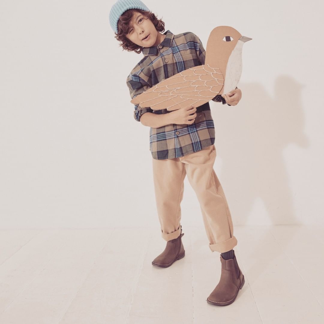 Boy in plaid shirt and beanie holds a cardboard bird in a bright studio, posing with a playful expression and wearing brown chelsea boots.