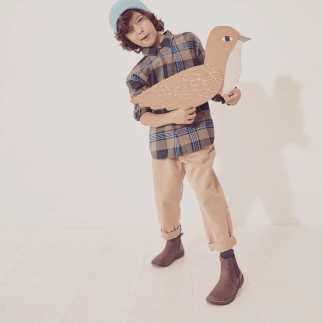 Boy in plaid shirt and beanie holds a cardboard bird in a bright studio, posing with a playful expression and wearing brown chelsea boots.