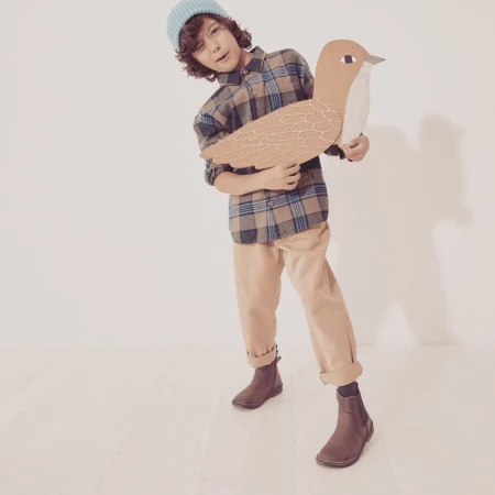 Boy in plaid shirt and beanie holds a cardboard bird in a bright studio, posing with a playful expression and wearing brown chelsea boots.
