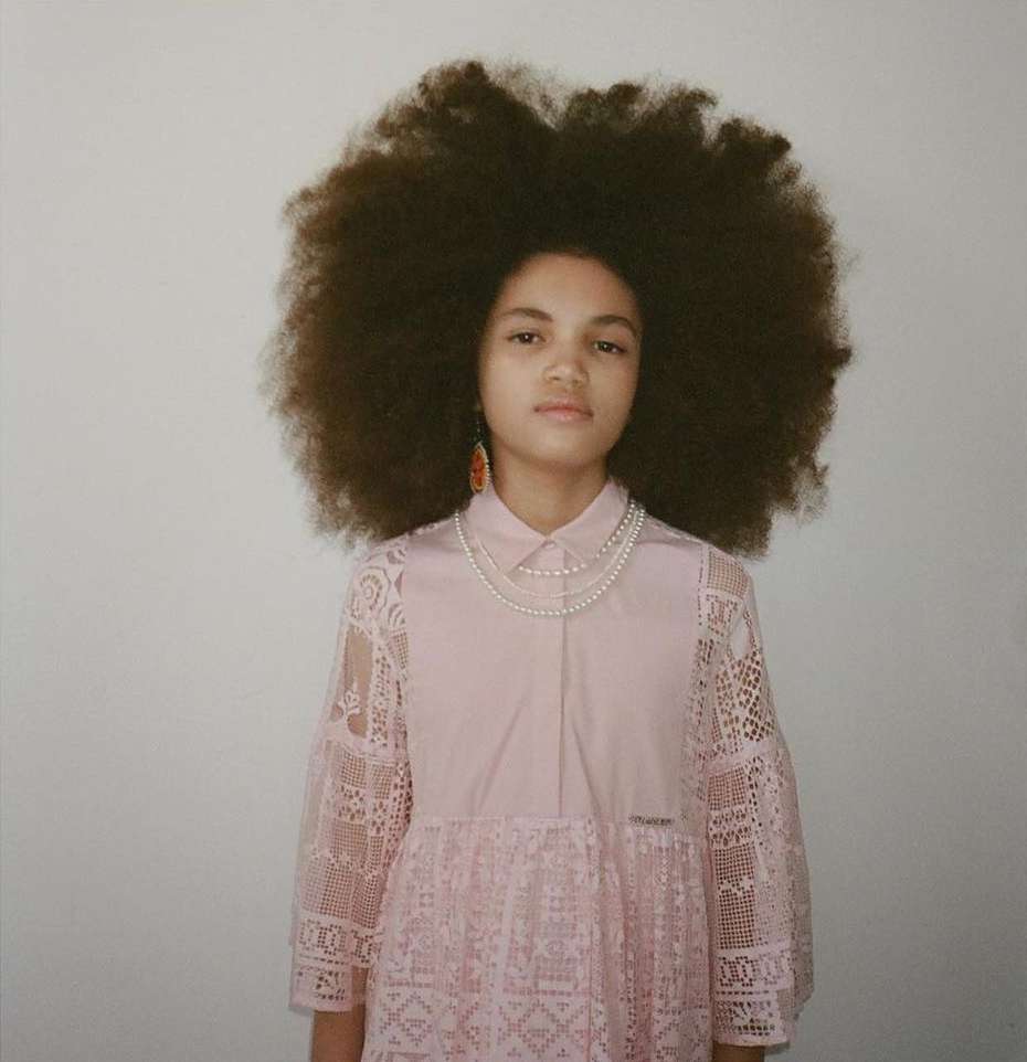 Young girl with a large afro hairstyle poses in a studio wearing a pink, lace-trimmed dress and pearl necklaces against a neutral background.