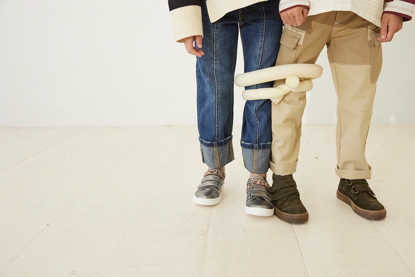 Two stylish children in casual outfits pose indoors. One wears jeans and the other wears khaki pants with neutral tops and sneakers.