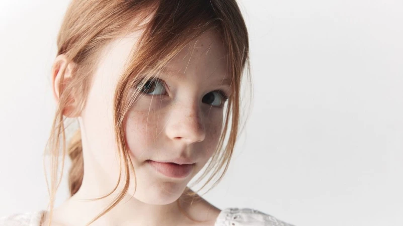 Close-up portrait of a young child with long red hair and soft freckles wearing a white embroidered top against a plain light background
