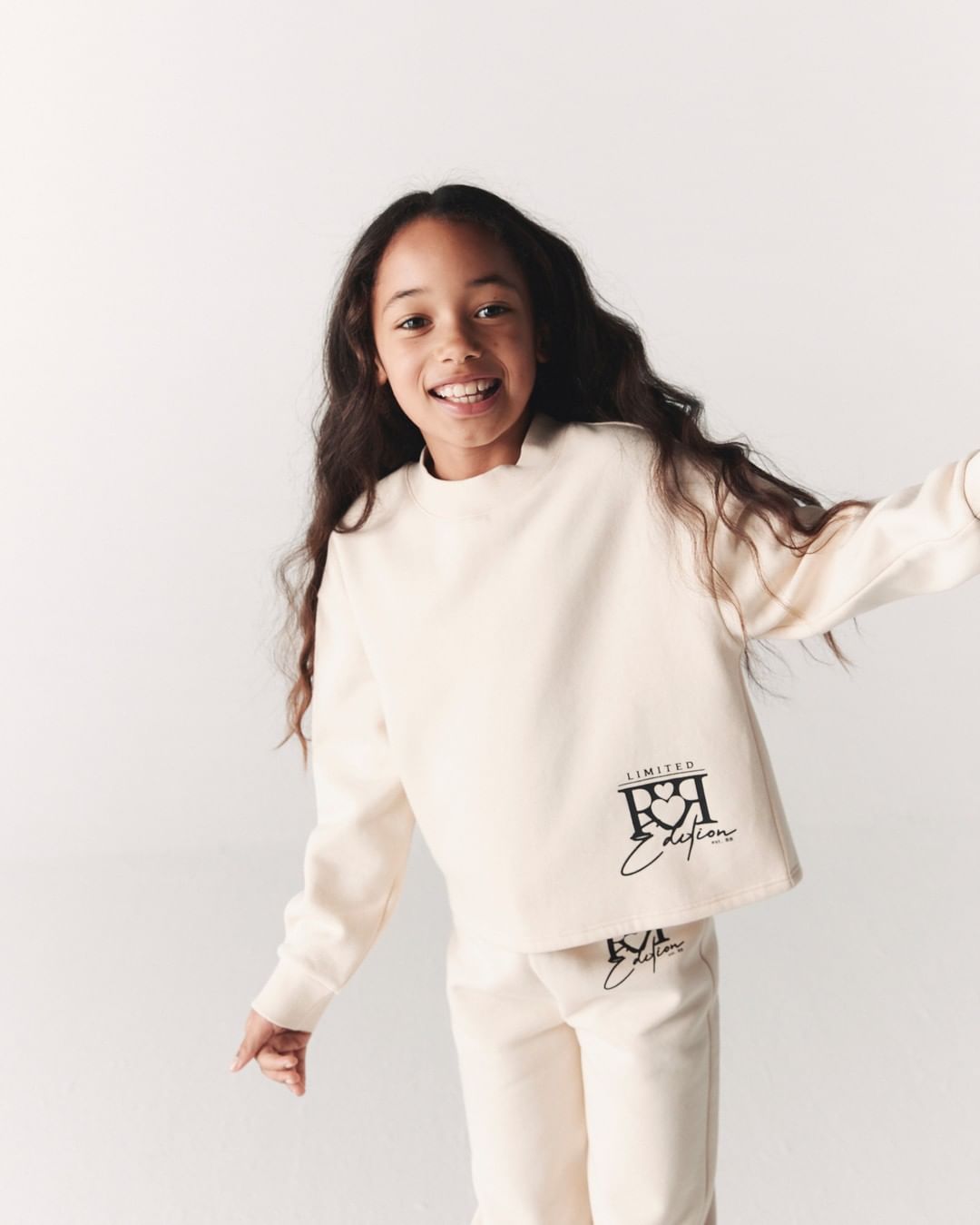 A smiling girl in a cream-colored sweatshirt and pants poses in a studio against a white backdrop, showcasing the fashion apparel.