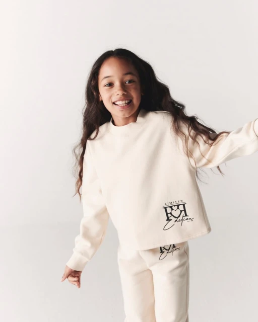 A smiling girl in a cream-colored sweatshirt and pants poses in a studio against a white backdrop, showcasing the fashion apparel.