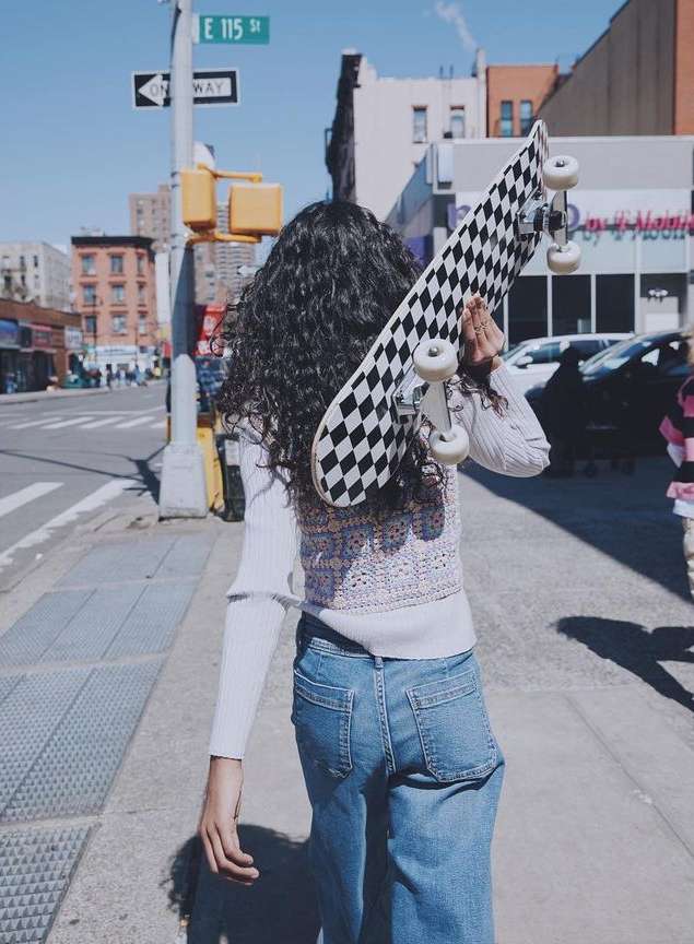 Young woman with curly hair walks along a city sidewalk carrying a black-and-white checkered skateboard over her shoulder.