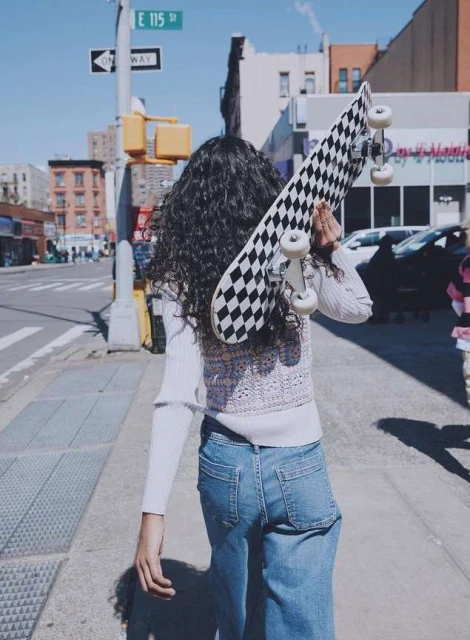 Young woman with curly hair walks along a city sidewalk carrying a black-and-white checkered skateboard over her shoulder.