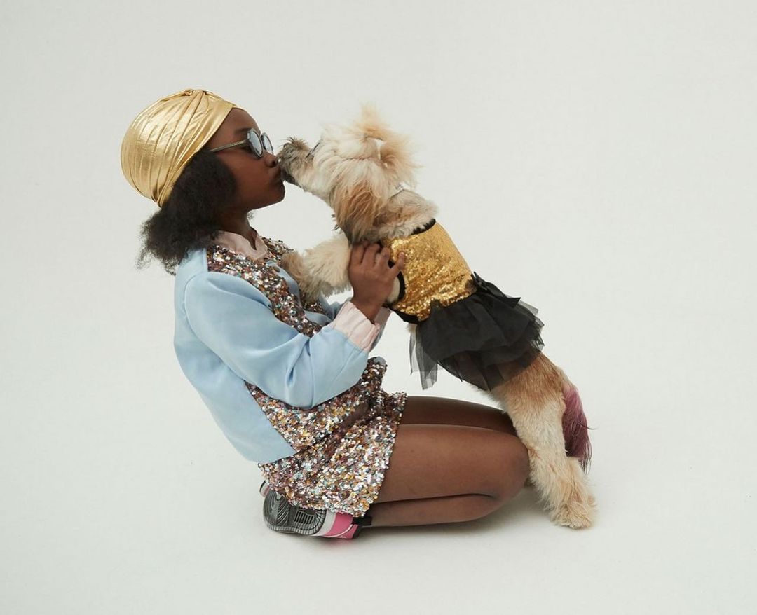 Young girl wearing a gold turban kisses a small dog in a gold sequin top and black tutu against a neutral studio backdrop.