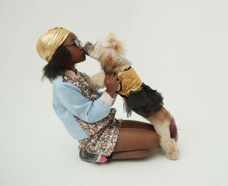 Young girl wearing a gold turban kisses a small dog in a gold sequin top and black tutu against a neutral studio backdrop.