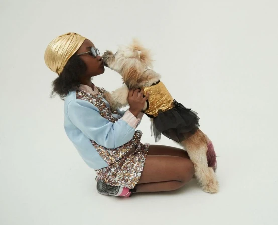 Young girl wearing a gold turban kisses a small dog in a gold sequin top and black tutu against a neutral studio backdrop.