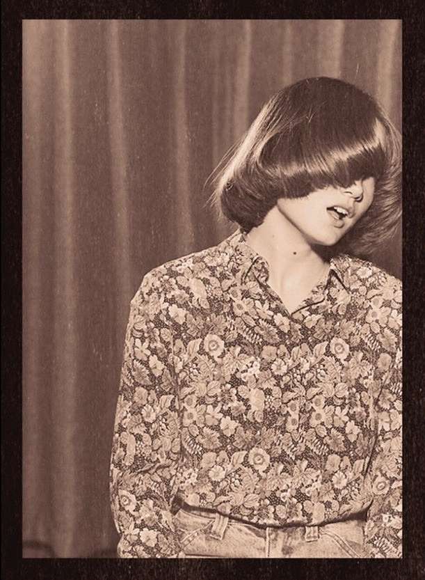 Portrait of a young adult woman with a floral shirt and bangs covering her eyes, in front of a draped backdrop, vintage photo.