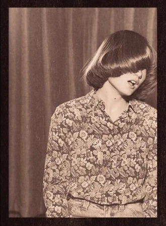 Portrait of a young adult woman with a floral shirt and bangs covering her eyes, in front of a draped backdrop, vintage photo.