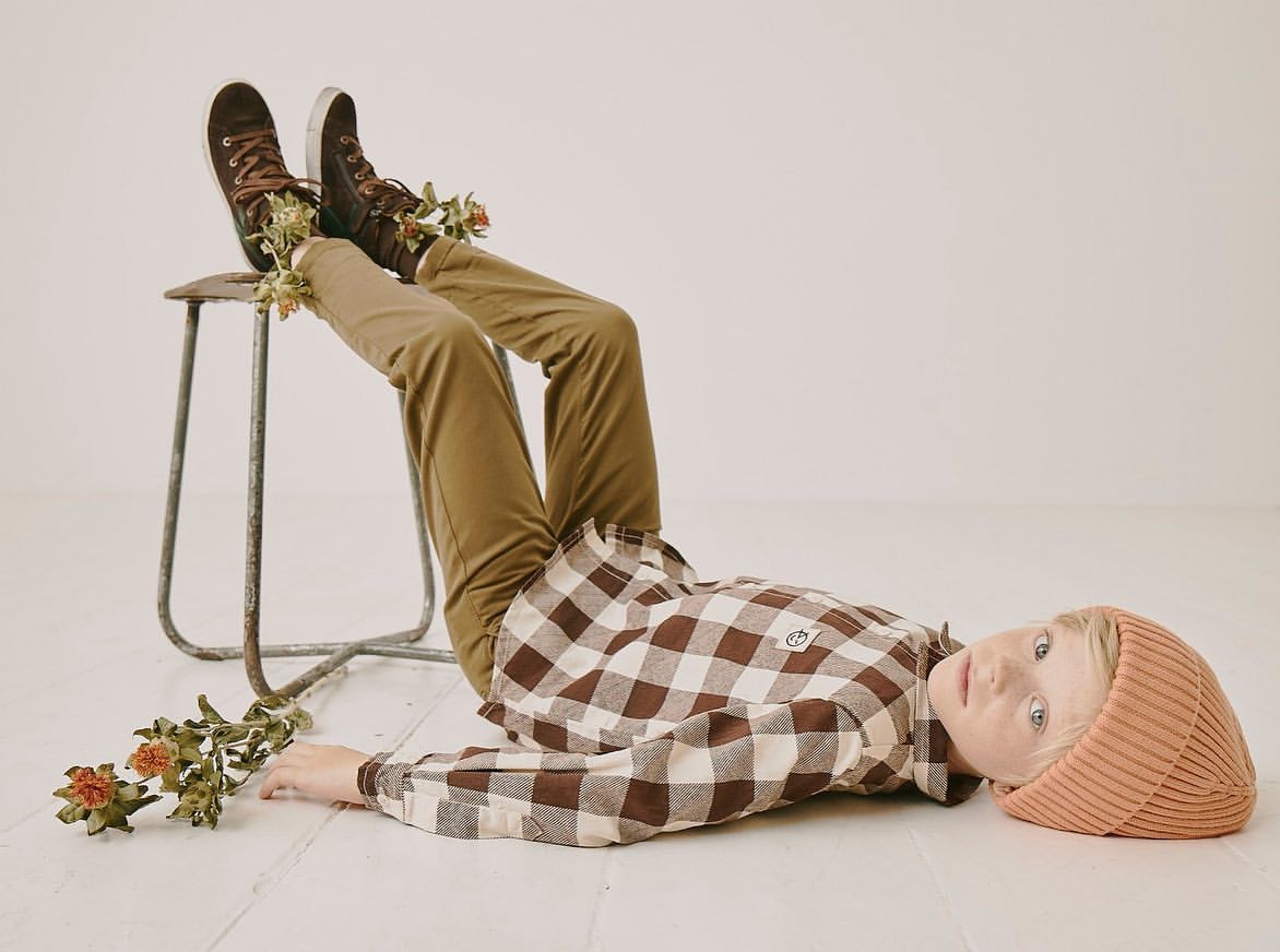 Boy lies on floor wearing brown and white checkered shirt and beanie. He has brown sneakers on a stool, and dried flowers.