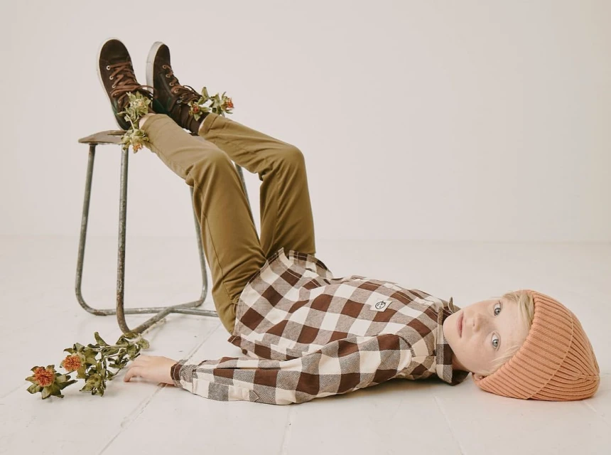 Boy lies on floor wearing brown and white checkered shirt and beanie. He has brown sneakers on a stool, and dried flowers.