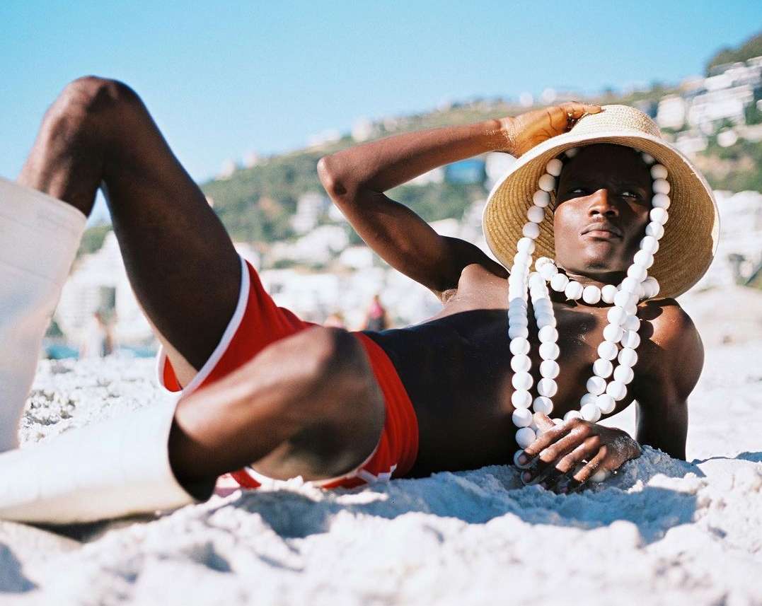 A single Black male on a sandy beach wearing red shorts, white boots, a straw hat, and a white beaded necklace against a sunny sky.