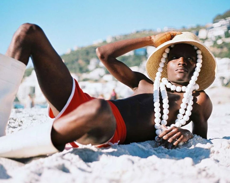 A single Black male on a sandy beach wearing red shorts, white boots, a straw hat, and a white beaded necklace against a sunny sky.