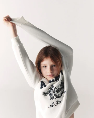 Young model stretching arms up while wearing an off-white sweatshirt against a seamless white background.