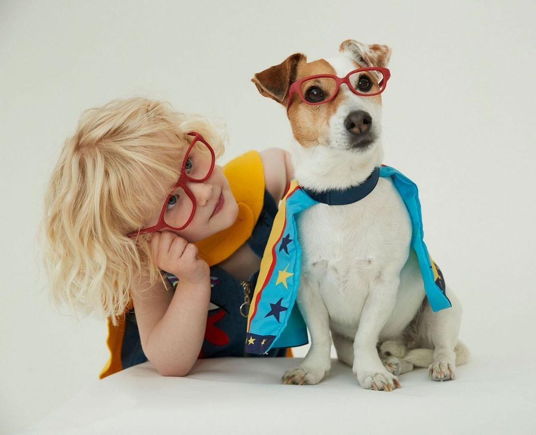 Adorable scene featuring a blonde child with glasses and a Jack Russell Terrier wearing glasses and a cape against a white background.