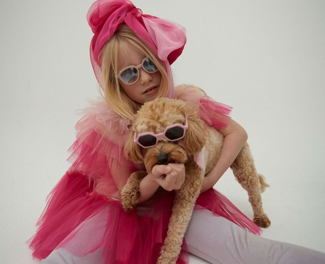 Young girl in pink tulle outfit and sunglasses holding a poodle, also wearing sunglasses, on a white background, studio lighting.
