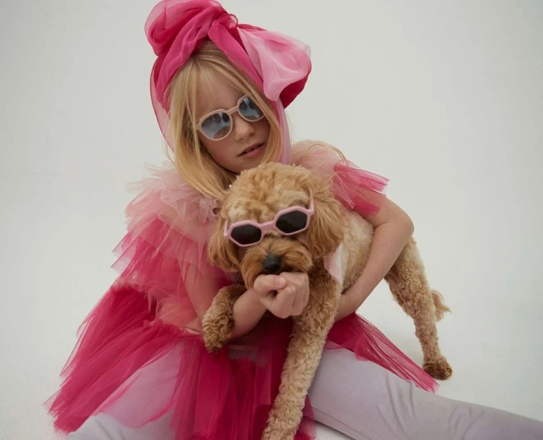 Young girl in pink tulle outfit and sunglasses holding a poodle, also wearing sunglasses, on a white background, studio lighting.