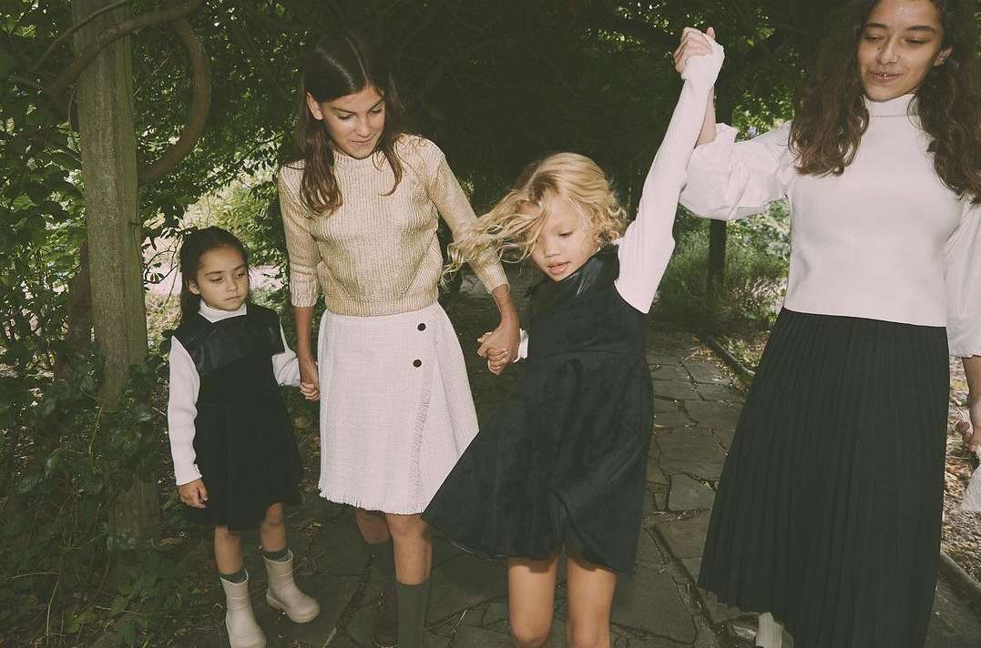 Four young girls walk holding hands on a stone path surrounded by lush greenery, wearing dresses, skirts and sweaters in an outdoor setting.
