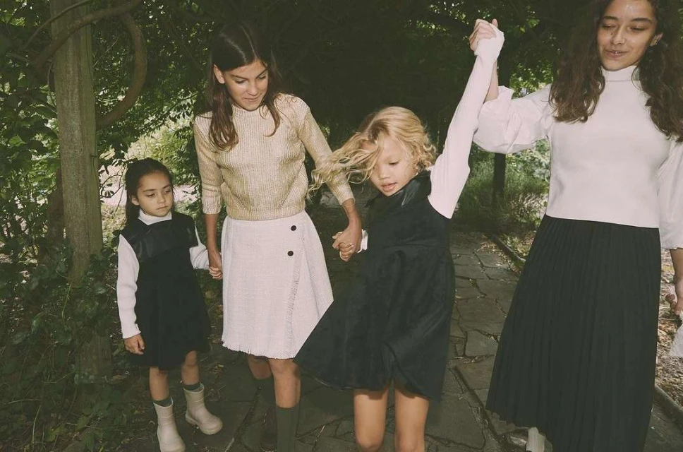 Four young girls walk holding hands on a stone path surrounded by lush greenery, wearing dresses, skirts and sweaters in an outdoor setting.