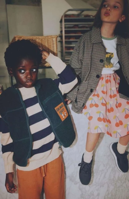Two young children in stylish outerwear, one Black child in a vest and striped shirt, the other a White child in a jacket and skirt.