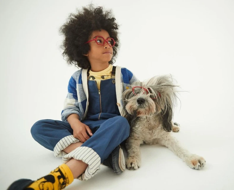 A young child with an afro and red glasses sits next to a fluffy grey dog also wearing red glasses in a bright, playful photo studio setting.