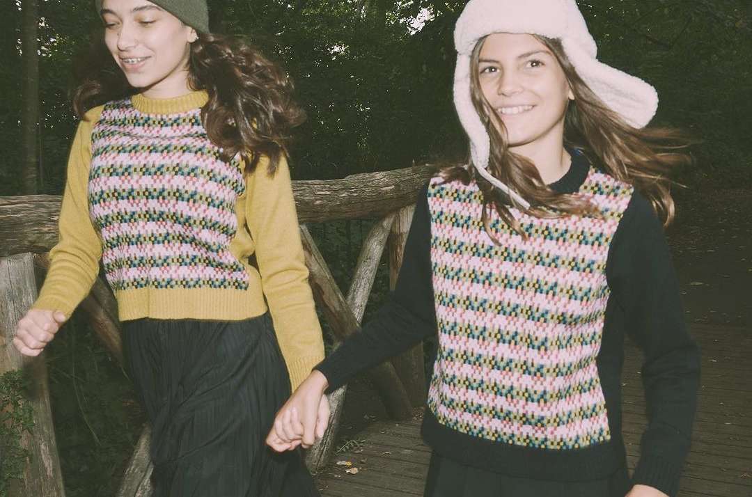 Two young girls in sweaters and skirts are walking across a wooden bridge in a forest; one has on a hat, smiling, outdoor setting.